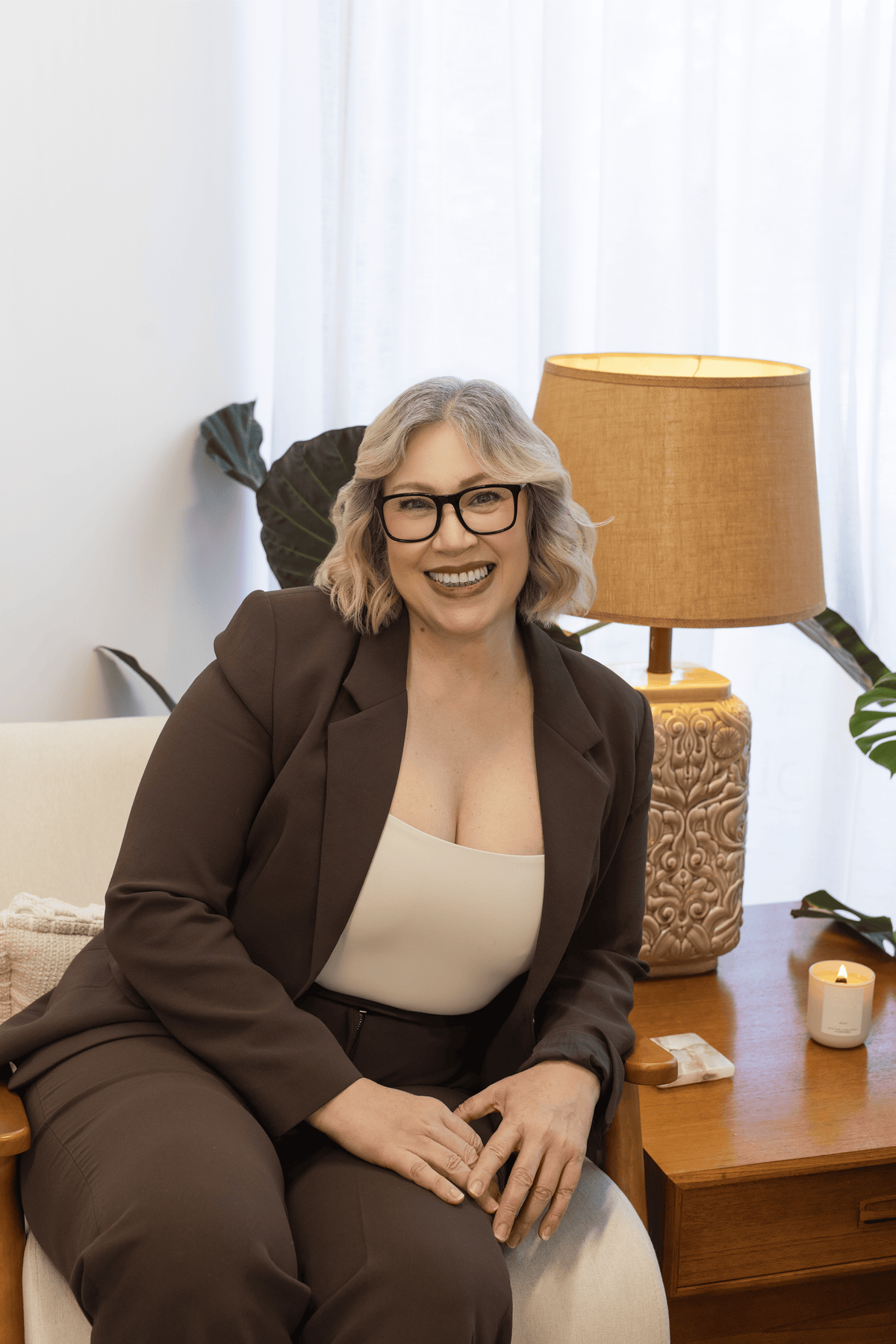 A smiling woman with short gray hair and glasses sits in a chair wearing a brown tailored blazer and matching pants over a cream top. She is posed indoors beside a wooden table with a lamp, candle, and plant in a bright neutral room.