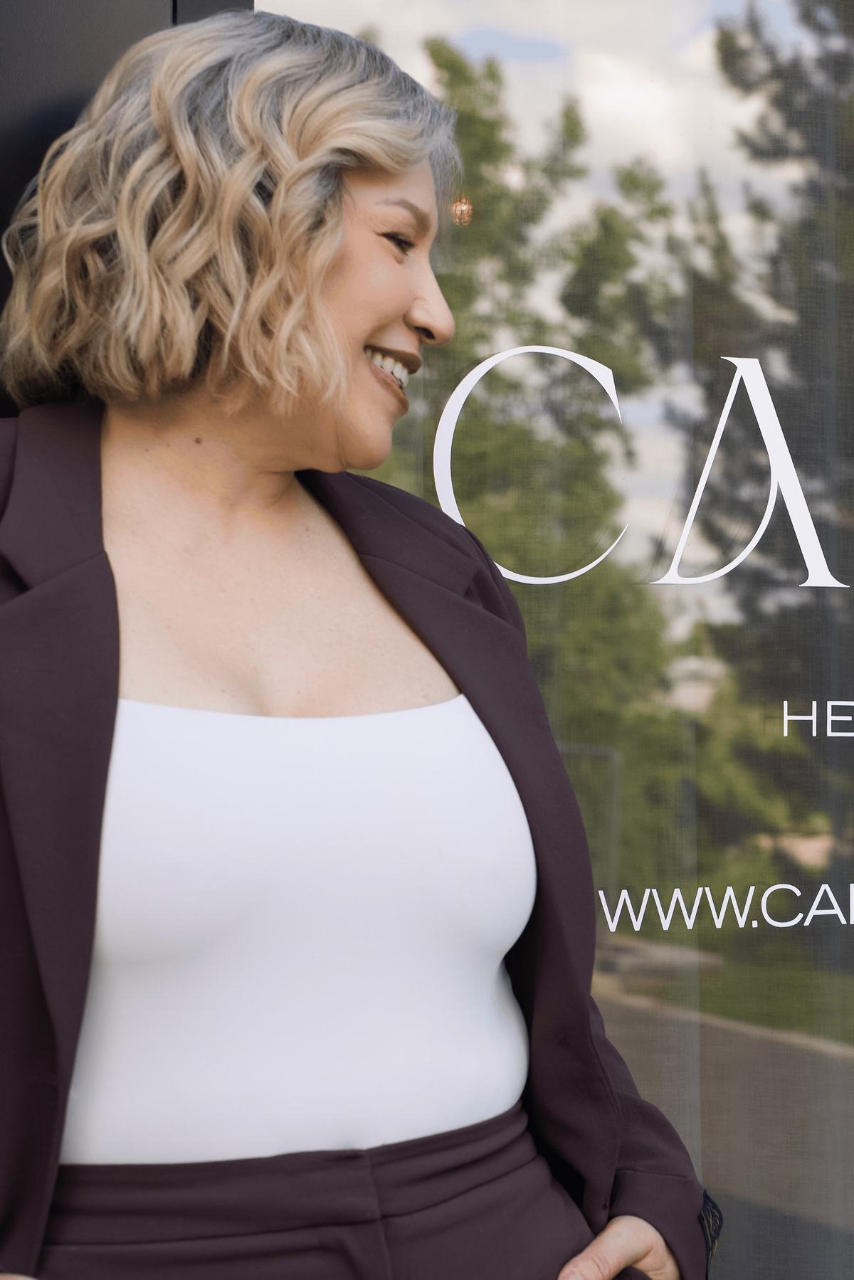A woman with short blonde hair stands in front of a glass storefront window with the text 'CAPITAL HEAD SPA & HAIR' visible. She is wearing a dark plum blazer and matching pants over a white top, with her hands in her pockets and smiling to the side.